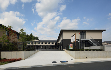 介護施設の建物外観。モダンな平屋造りで、中庭を囲む構造。白い舗装と木製デッキが見える。
